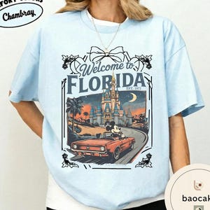 May include: Light blue t-shirt with a graphic that says "Welcome to Florida" featuring a vintage car with Mickey Mouse driving past a castle. The shirt has the text "Comfort Colors Chambray" on the top left.