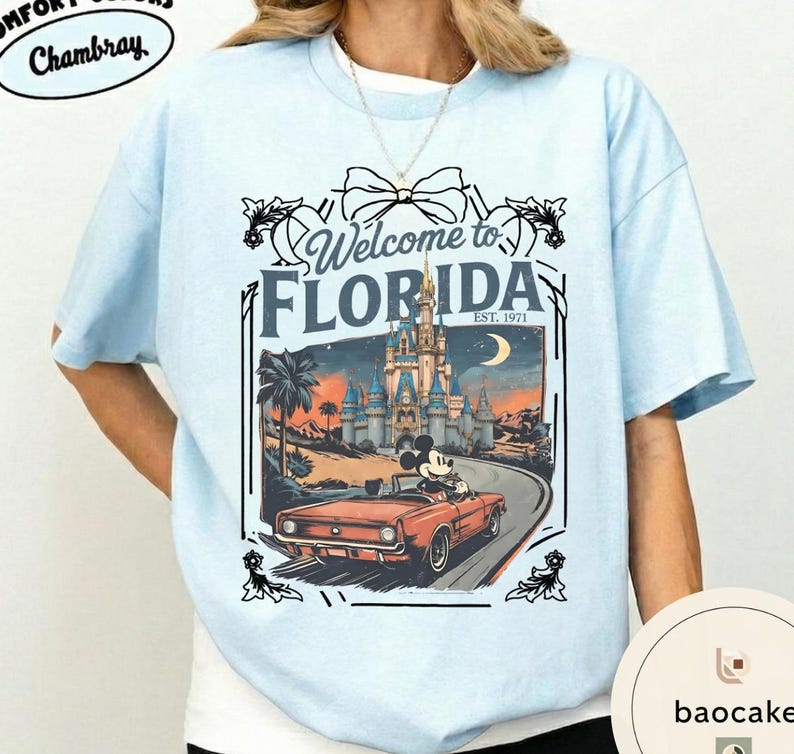 May include: Light blue t-shirt with a graphic that says "Welcome to Florida" featuring a vintage car with Mickey Mouse driving past a castle. The shirt has the text "Comfort Colors Chambray" on the top left.