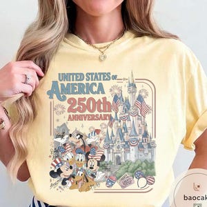 May include: Yellow t-shirt featuring a graphic celebrating the 250th anniversary of the United States of America. The design includes a castle, fireworks, and Disney characters like Mickey Mouse and Donald Duck.