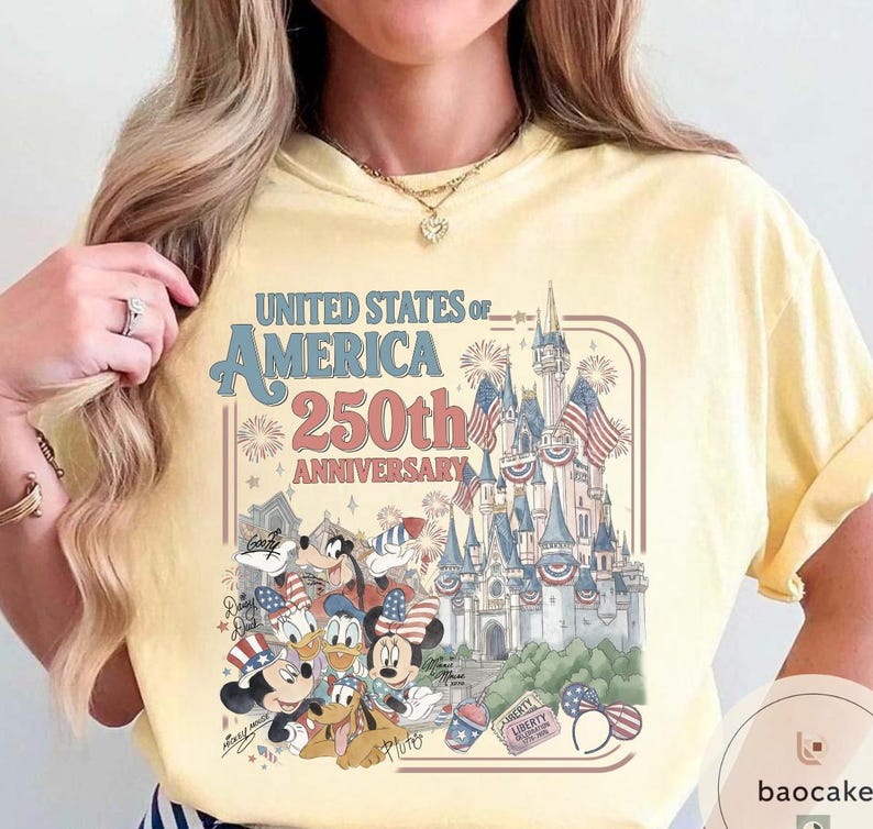 May include: Yellow t-shirt featuring a graphic celebrating the 250th anniversary of the United States of America. The design includes a castle, fireworks, and Disney characters like Mickey Mouse and Donald Duck.