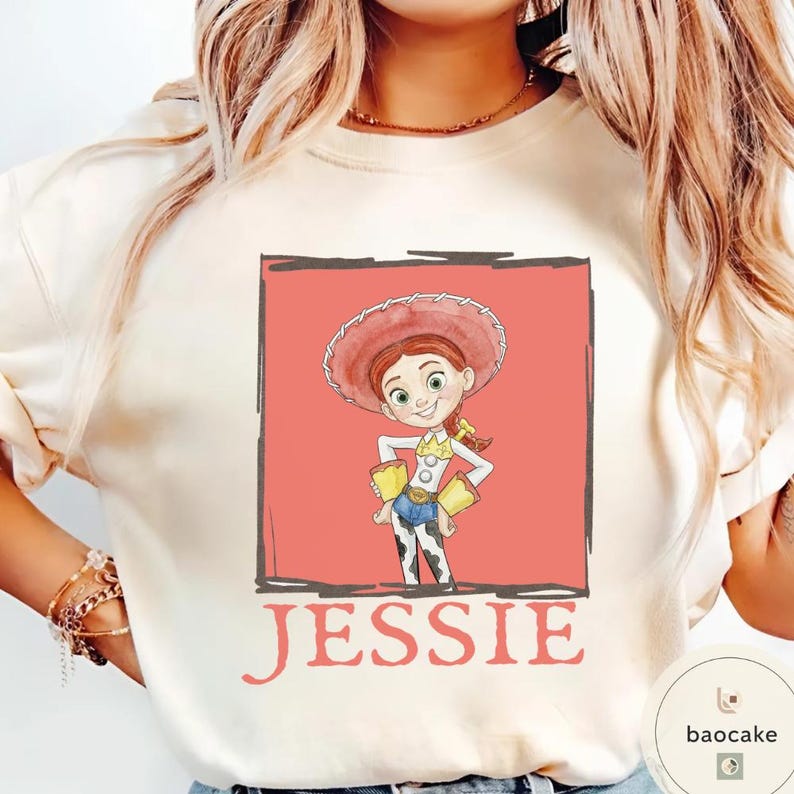 K&ouml;nnte beinhalten: Ein cremefarbenes T-Shirt mit einer rechteckigen Grafik der Figur Jessie aus Toy Story. Die Illustration zeigt Jessie mit einem roten Cowgirl-Hut, einem wei&szlig;en Hemd mit gelben Manschetten und einer Hose mit Kuhfleckenmuster. Der Name JESSIE ist darunter aufgedruckt.