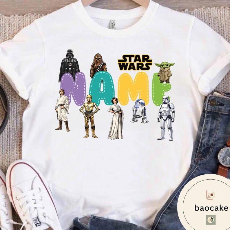 May include: White t-shirt featuring Star Wars characters and the text "NAME" in colorful bubble letters. The design includes Darth Vader, Chewbacca, Luke Skywalker, C-3PO, Princess Leia, R2-D2, a Stormtrooper, and Grogu.