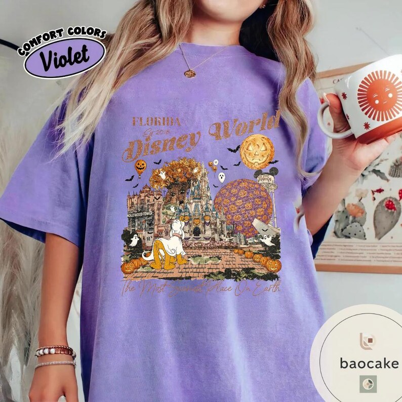 Op de afbeelding: Violet Comfort Colors T-shirt met een Halloween-thema Florida Disney World-afbeelding. Het ontwerp bevat pompoenen, geesten en de tekst "Florida Disney World 2018". Het shirt heeft een relaxte pasvorm.