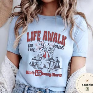 May include: Light blue t-shirt featuring a vintage-style graphic. The design includes the text "LIFE AWALK IN THE PARK" in red, with a cartoon character and a castle illustration. The words "Walt Disney World" are also printed on the shirt.