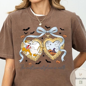 May include: Brown t-shirt with a Halloween graphic. The design features a heart-shaped locket with cartoon characters, bats, spiders, and pumpkins. The text "The most scariest place on earth" is printed below the locket.