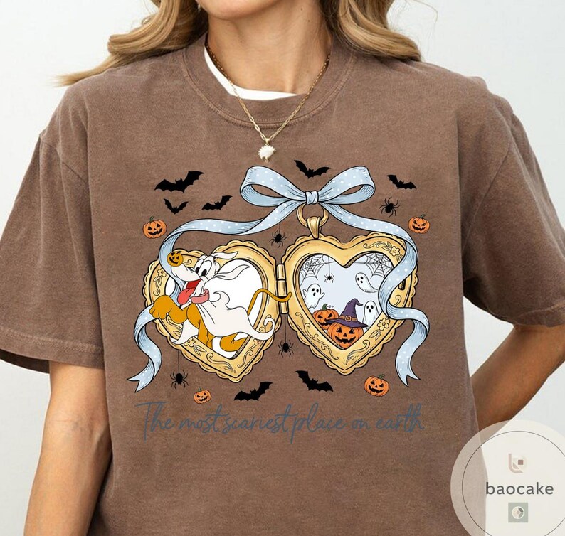 May include: Brown t-shirt with a Halloween graphic. The design features a heart-shaped locket with cartoon characters, bats, spiders, and pumpkins. The text "The most scariest place on earth" is printed below the locket.