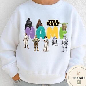 May include: White sweatshirt featuring Star Wars characters and the word "NAME" in colorful letters. Characters include Darth Vader, Chewbacca, Yoda, and others. The Star Wars logo is also visible.