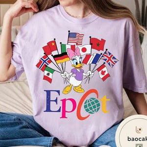May include: A lilac t-shirt with a Daisy Duck graphic holding flags of different nations. The word "Epcot" is printed in colourful letters below the character, with a globe graphic. The shirt has a relaxed fit.