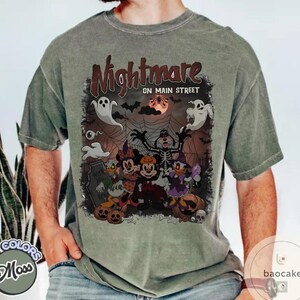 May include: A moss green t-shirt featuring a Halloween-themed graphic with the text "Nightmare on Main Street." The design includes cartoon characters dressed in Halloween costumes, ghosts, bats, and a full moon.