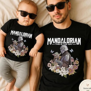 Puede incluir: Camisetas negras con el texto "MANDALORIAN - THIS IS THE WAY" en blanco. Las camisetas presentan un gr&aacute;fico del Mandaloriano y Grogu rodeados de flores y mariposas. Una camiseta es para un adulto, la otra para un beb&eacute;.