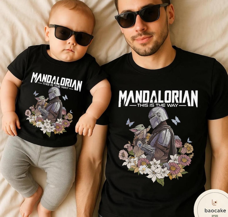 Puede incluir: Camisetas negras con el texto "MANDALORIAN - THIS IS THE WAY" en blanco. Las camisetas presentan un gr&aacute;fico del Mandaloriano y Grogu rodeados de flores y mariposas. Una camiseta es para un adulto, la otra para un beb&eacute;.