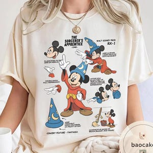 May include: Cream-colored t-shirt featuring a graphic of Mickey Mouse in his Sorcerer's Apprentice attire. The design includes illustrations and text detailing Mickey's features and the film's elements. The text includes "The Sorcerer's Apprentice - 1940".