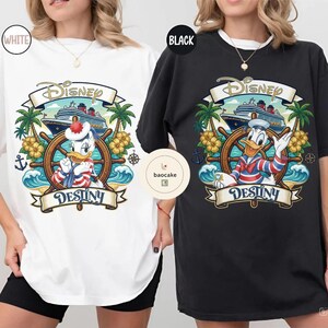 May include: White and black t-shirts featuring a Disney-themed graphic. The design includes Donald Duck at a ship's wheel, a cruise ship, palm trees, and the word "Destiny" on a banner. The Disney logo is at the top.