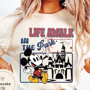 May include: Cream-coloured t-shirt featuring Mickey Mouse pointing towards a castle silhouette. The shirt has the text "LIFE AWALK IN THE Park" in red and blue, with fireworks and stars.
