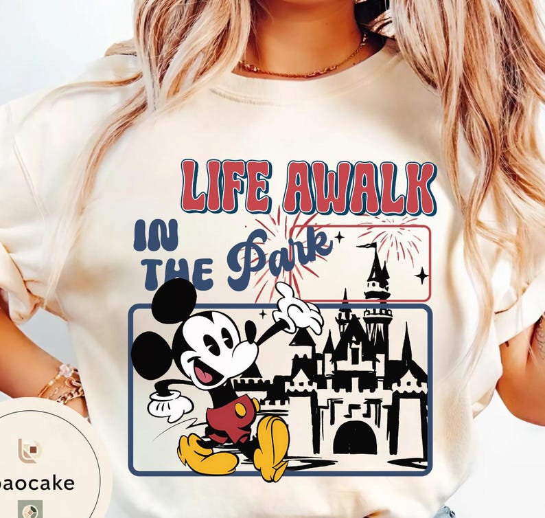 May include: Cream-coloured t-shirt featuring Mickey Mouse pointing towards a castle silhouette. The shirt has the text "LIFE AWALK IN THE Park" in red and blue, with fireworks and stars.