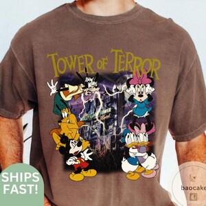 May include: Brown t-shirt featuring a graphic of Disney characters, including Mickey Mouse, Minnie Mouse, Goofy, Donald Duck, and Pluto, with the text "Tower of Terror". The design includes a purple lightning effect and a building.