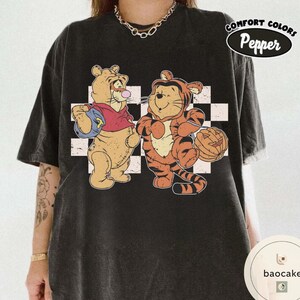 May include: Black Comfort Colors t-shirt featuring cartoon characters Winnie the Pooh and Tigger. Winnie the Pooh is wearing a red shirt and holding a blue bag. Tigger is dressed in an orange tiger costume and holding a jack-o'-lantern. The shirt has a checkered background.