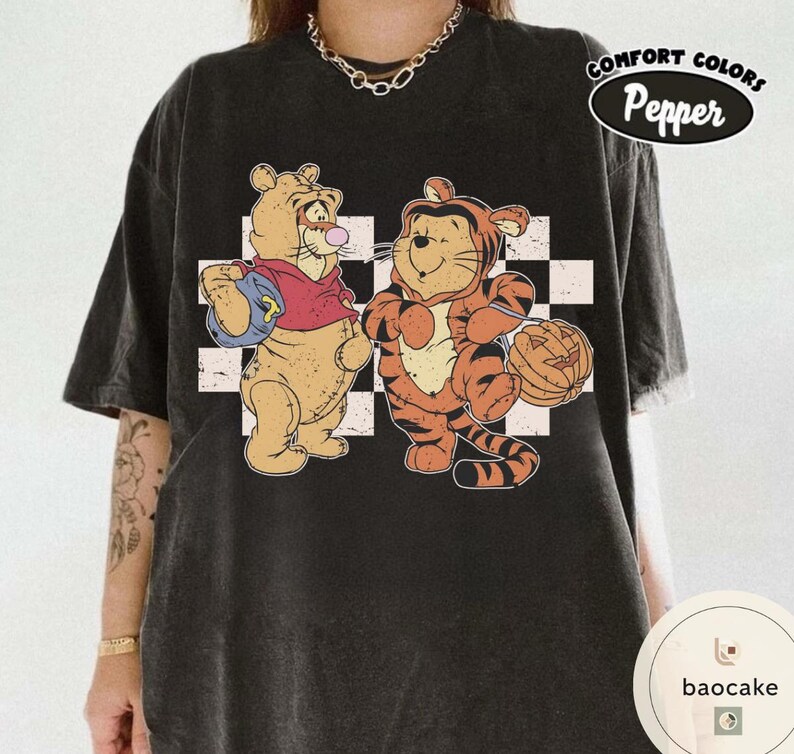 May include: Black Comfort Colors t-shirt featuring cartoon characters Winnie the Pooh and Tigger. Winnie the Pooh is wearing a red shirt and holding a blue bag. Tigger is dressed in an orange tiger costume and holding a jack-o'-lantern. The shirt has a checkered background.