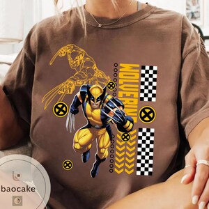 May include: Brown t-shirt featuring a Wolverine graphic in yellow and blue. The design includes a detailed illustration of Wolverine in action, the word "Wolverine", and a checkered pattern. The shirt has a crew neck and short sleeves.