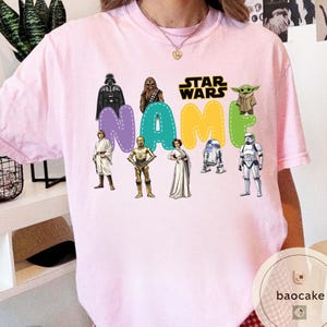 May include: A light pink t-shirt featuring a Star Wars design. The design includes the Star Wars logo, the word "NAME" in colorful letters, and various Star Wars characters such as Darth Vader, Chewbacca, and R2-D2.