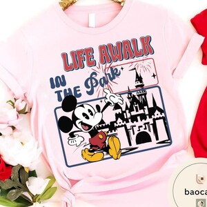 May include: Pale pink t-shirt with a graphic of Mickey Mouse and the text "LIFE A WALK IN THE Park". The design includes a castle, fireworks, and a blue border. Mickey is in red shorts and yellow shoes.