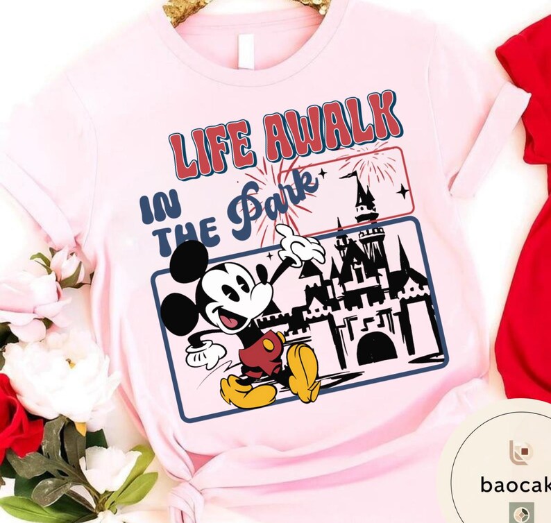 May include: Pale pink t-shirt with a graphic of Mickey Mouse and the text "LIFE A WALK IN THE Park". The design includes a castle, fireworks, and a blue border. Mickey is in red shorts and yellow shoes.
