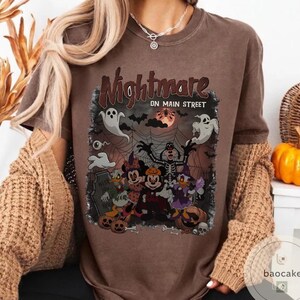 May include: Brown t-shirt with a Halloween-themed graphic. The graphic features cartoon characters dressed in Halloween costumes, ghosts, bats, and the text "Nightmare on Main Street". The design includes a full moon and spiderwebs.