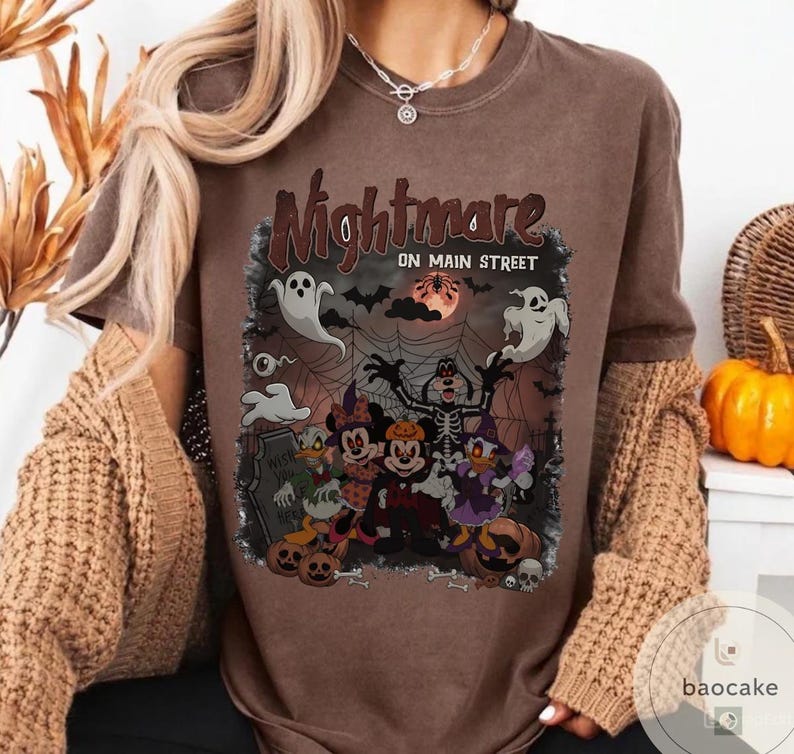 May include: Brown t-shirt with a Halloween-themed graphic. The graphic features cartoon characters dressed in Halloween costumes, ghosts, bats, and the text "Nightmare on Main Street". The design includes a full moon and spiderwebs.