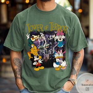 May include: Green t-shirt featuring a graphic of Mickey Mouse, Minnie Mouse, Donald Duck, and Goofy in front of the Tower of Terror. The text "Tower of Terror" is printed in gold across the top of the shirt.