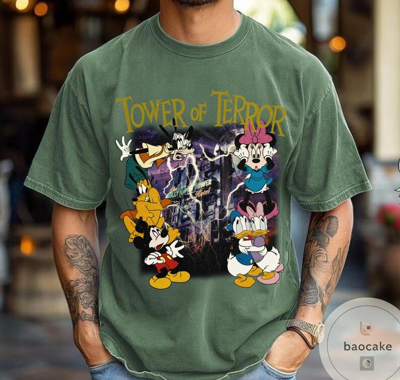May include: Green t-shirt featuring a graphic of Mickey Mouse, Minnie Mouse, Donald Duck, and Goofy in front of the Tower of Terror. The text "Tower of Terror" is printed in gold across the top of the shirt.