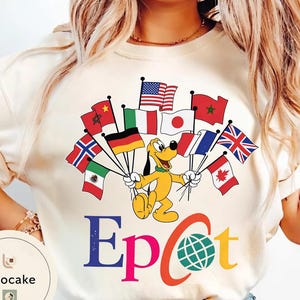 May include: Cream-coloured t-shirt with a cartoon Pluto holding flags from various countries, including the United States, Japan, Italy, and the United Kingdom. The word "Epcot" is printed in colourful letters below the cartoon.