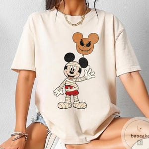 May include: Cream-colored t-shirt featuring a cartoon Mickey Mouse dressed as a mummy, holding a brown balloon with a jack-o'-lantern face. Mickey is wearing bandages and red shorts. The shirt has a round neck and short sleeves.