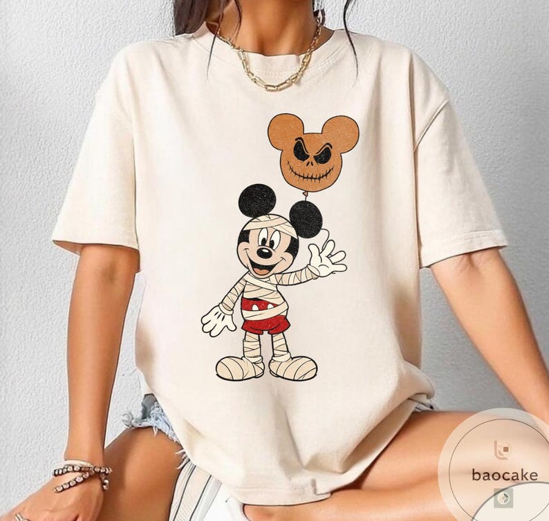 May include: Cream-colored t-shirt featuring a cartoon Mickey Mouse dressed as a mummy, holding a brown balloon with a jack-o'-lantern face. Mickey is wearing bandages and red shorts. The shirt has a round neck and short sleeves.