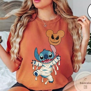 May include: Orange-colored t-shirt featuring a Halloween-themed graphic. The design includes a blue alien character wrapped in bandages, a Mickey Mouse-shaped jack-o'-lantern balloon, and the character's name. The shirt is a comfortable, casual style.