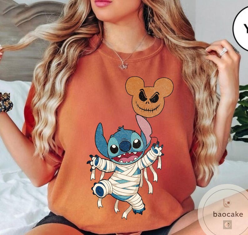 May include: Orange-colored t-shirt featuring a Halloween-themed graphic. The design includes a blue alien character wrapped in bandages, a Mickey Mouse-shaped jack-o'-lantern balloon, and the character's name. The shirt is a comfortable, casual style.