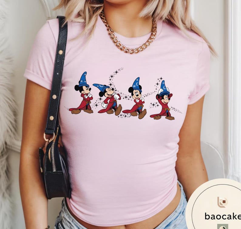 May include: A light pink t-shirt featuring a graphic of Mickey Mouse in a sorcerer's outfit. The cartoon characters are depicted in a row, wearing blue wizard hats and red robes, with a starry design.