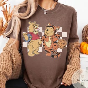 May include: Brown t-shirt featuring a cartoon illustration of Winnie the Pooh and Tigger dressed for Halloween. Winnie the Pooh is holding a football, and Tigger is holding a jack-o'-lantern. The background has a checkered pattern.