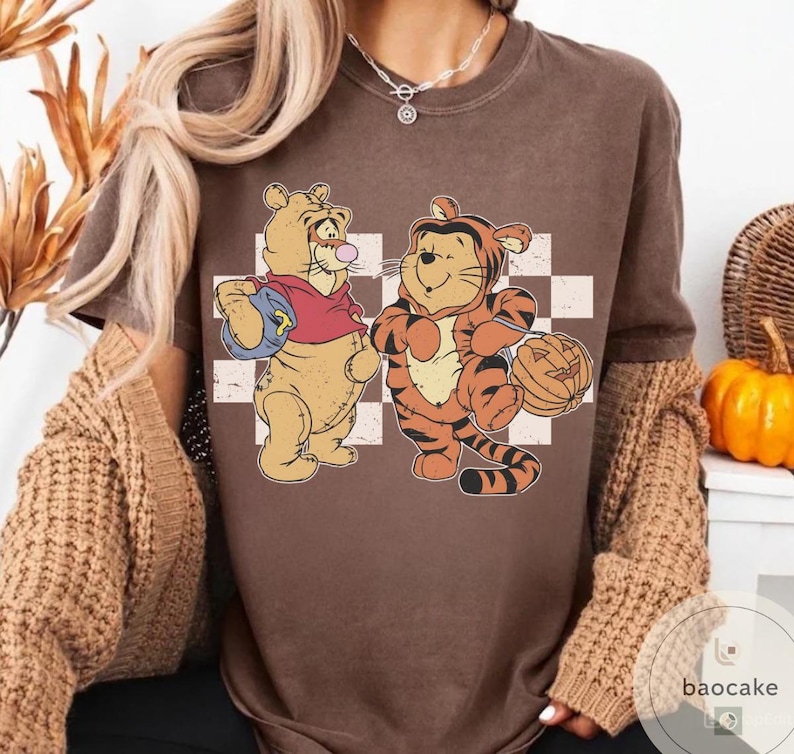 May include: Brown t-shirt featuring a cartoon illustration of Winnie the Pooh and Tigger dressed for Halloween. Winnie the Pooh is holding a football, and Tigger is holding a jack-o'-lantern. The background has a checkered pattern.