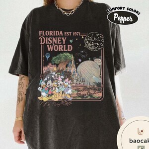 May include: A charcoal black short-sleeve t-shirt featuring a graphic print with the text FLORIDA EST 1971 DISNEY WORLD and The Most Magical Place On Earth. The design includes illustrations of Disney characters, the castle, and Epcot.