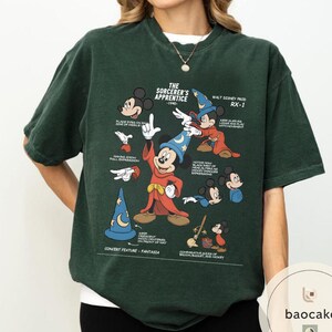 May include: Dark green t-shirt featuring a graphic of Mickey Mouse from "The Sorcerer's Apprentice." The design includes illustrations of Mickey in various poses, a wizard hat, and text. The shirt has short sleeves and a crew neck.