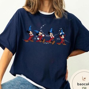 May include: Navy blue t-shirt featuring a graphic of Mickey Mouse in a wizard costume. The design shows Mickey Mouse in a red robe and blue wizard hat with a crescent moon and stars. The shirt has a white crew neck collar.