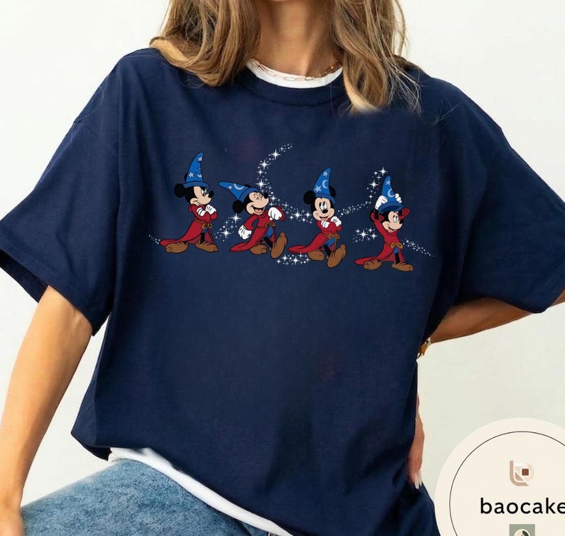 May include: Navy blue t-shirt featuring a graphic of Mickey Mouse in a wizard costume. The design shows Mickey Mouse in a red robe and blue wizard hat with a crescent moon and stars. The shirt has a white crew neck collar.