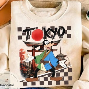 Puede incluir: Sudadera crema con un dise&ntilde;o de Goofy con vestimenta japonesa tradicional, con la palabra "TOKYO" arriba. El dise&ntilde;o incluye un sol rojo, una puerta japonesa y una pagoda.