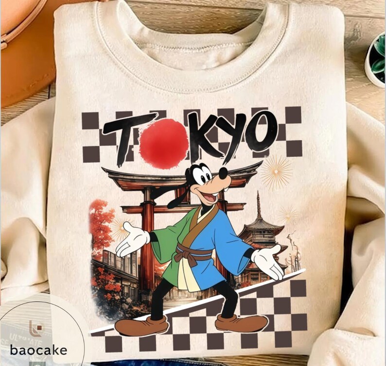 Puede incluir: Sudadera crema con un dise&ntilde;o de Goofy con vestimenta japonesa tradicional, con la palabra "TOKYO" arriba. El dise&ntilde;o incluye un sol rojo, una puerta japonesa y una pagoda.