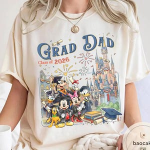 May include: Cream-coloured t-shirt with a colourful graphic featuring Disney characters and the text "Grad Dad Class of 2026." The design includes Mickey Mouse, Minnie Mouse, Donald Duck, Goofy, and Pluto, with a castle backdrop.