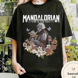 Puede incluir: Camiseta negra con un dise&ntilde;o de The Mandalorian y Grogu, rodeados de flores y mariposas. El texto "MANDALORIAN - THIS IS THE WAY" est&aacute; impreso en la parte superior. La camiseta es de color oscuro.