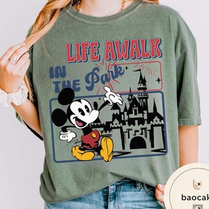 May include: Olive green t-shirt with a graphic of Mickey Mouse and the text "LIFE A WALK IN THE Park". Mickey is in red shorts and yellow shoes, with a castle and fireworks graphic.