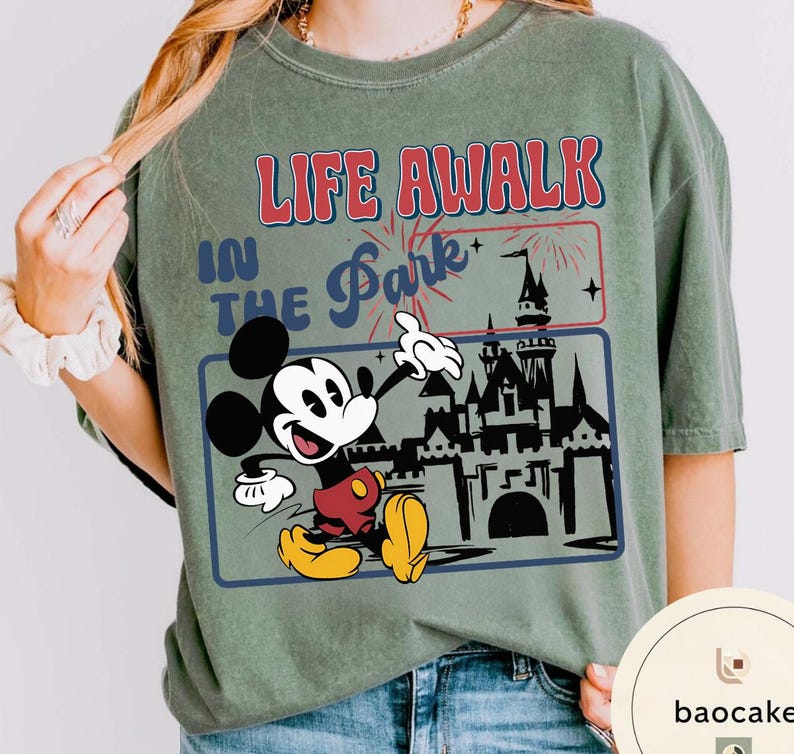 May include: Olive green t-shirt with a graphic of Mickey Mouse and the text "LIFE A WALK IN THE Park". Mickey is in red shorts and yellow shoes, with a castle and fireworks graphic.