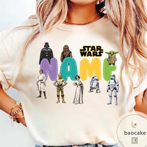 May include: Cream-colored Star Wars themed sweatshirt featuring the text "NAME" in colorful bubble letters. The design includes images of Darth Vader, Chewbacca, Yoda, and other characters from the Star Wars franchise.
