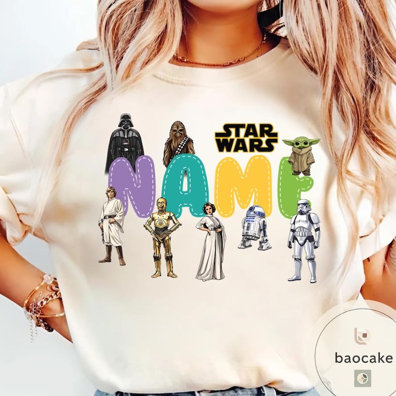 May include: Cream-colored Star Wars themed sweatshirt featuring the text "NAME" in colorful bubble letters. The design includes images of Darth Vader, Chewbacca, Yoda, and other characters from the Star Wars franchise.